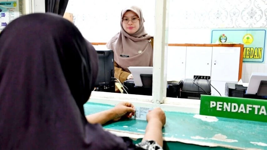 Seorang petugas wanita berjilbab melayani pasien di meja pendaftaran rumah sakit atau puskesmas.