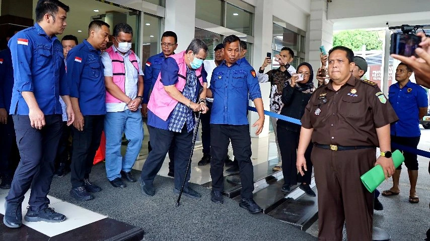 Dua pria paruh baya berompi tahanan pink dan kemeja kotak-kotak berjalan dibantu tongkat, dikawal petugas kejaksaan.