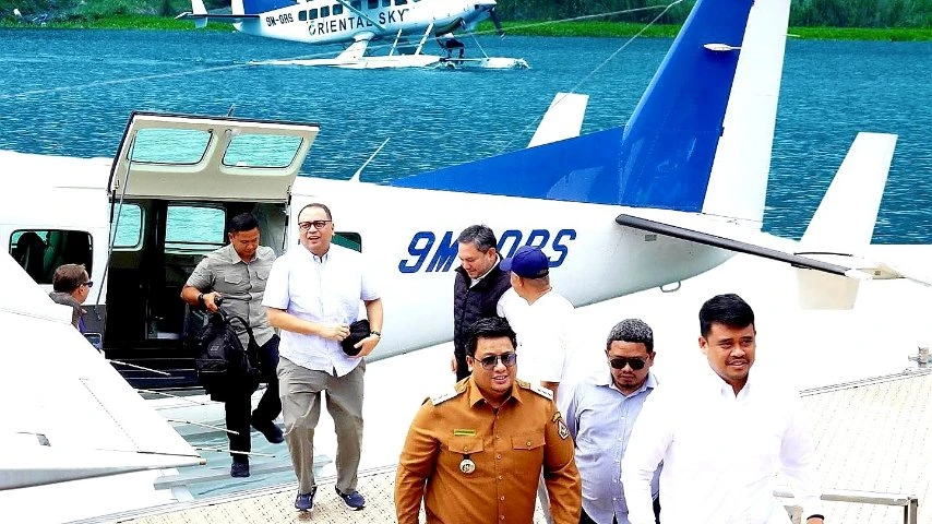 Gubernur Sumatera Utara Bobby Nasution mengenakan kemeja putih dan celana krem berjalan keluar dari pesawat amfibi di dermaga Danau Toba, dikelilingi oleh rombongan pejabat.