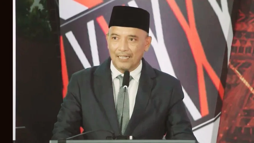 Pria paruh baya berkulit sawo matang mengenakan setelan jas biru gelap, kemeja putih, dasi abu-abu bermotif, dan peci hitam, sedang berbicara di depan mikrofon dengan latar belakang abstrak berwarna merah dan hitam.