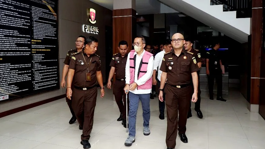 Sekelompok pria berseragam cokelat kejaksaan mengawal seorang pria berkacamata dengan rompi tahanan merah muda dan celana biru tua yang sedang berjalan tertatih menggunakan tongkat di dalam gedung kantor.