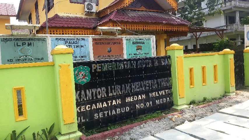 Kantor Lurah Helvetia Timur di Medan, dengan papan nama besar berwarna hitam dan kuning yang bertuliskan "PEMERINTAH KOTA MEDAN KANTOR LURAH HELVETIA TIMUR KECAMATAN MEDAN HELVETIA JL. SETIABUDI NO. 01 MEDAN," diapit oleh pagar tembok berwarna hijau limau dan kuning.