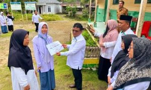 Seorang pria berkemeja putih menyerahkan sertifikat kepada siswi berhijab putih-ungu di luar ruangan. Di sekitar mereka ada beberapa orang dewasa dan siswi lain yang menyaksikan, dengan latar belakang bangunan sekolah berwarna hijau dan pepohonan.