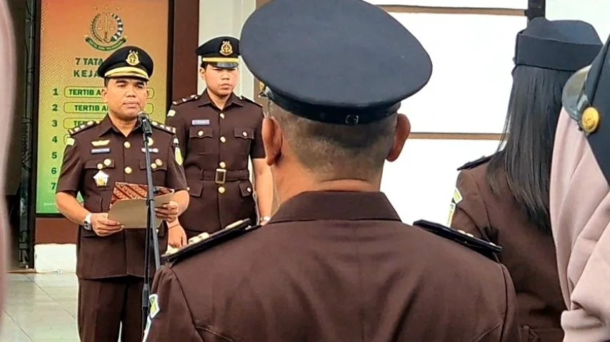 Pria berjas cokelat dengan peci hitam, diduga pejabat, sedang berbicara di depan sekelompok orang berseragam cokelat dan hitam di luar sebuah gedung.