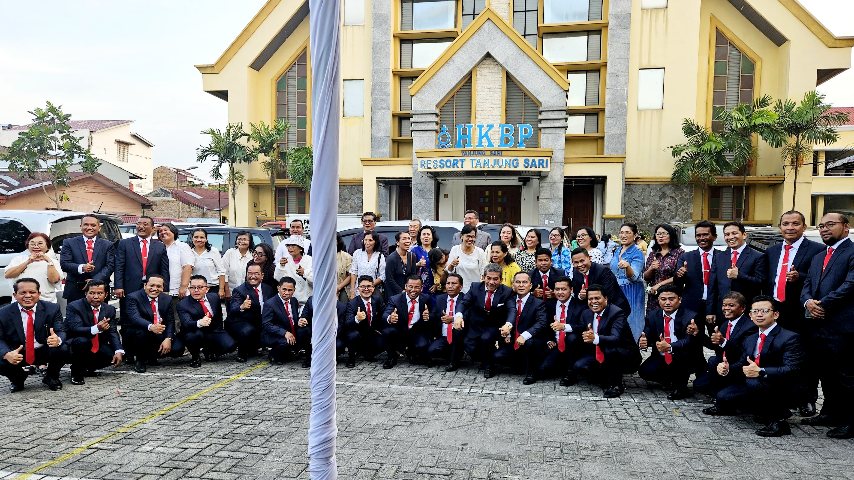 Sekelompok besar pria dan wanita berpose bersama di depan sebuah gereja. Para pria mengenakan jas hitam dan dasi merah, sementara para wanita mengenakan atasan putih.