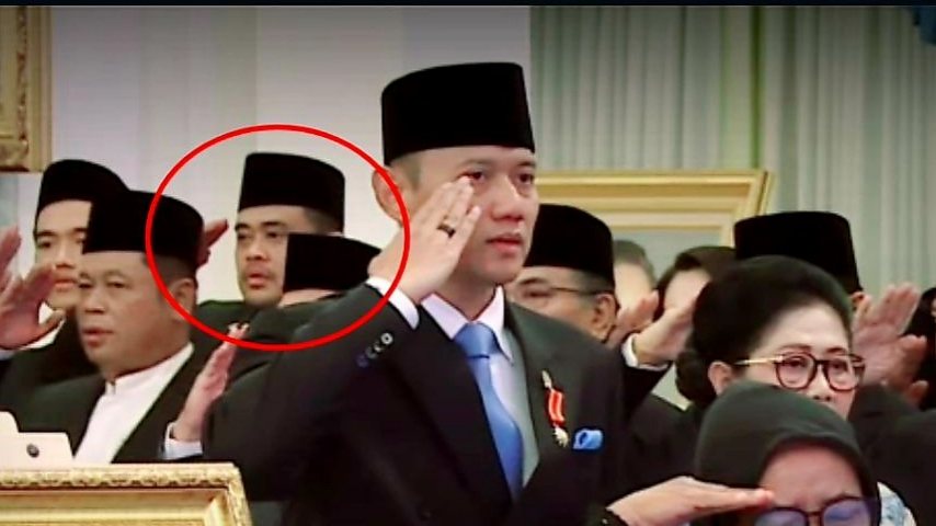Sekelompok orang berseragam jas hitam dan peci hitam sedang memberi hormat, dengan seorang pria di depan, di tengah, dan seorang wanita di sebelah kanan bawah. Ada bingkai foto di kiri bawah.