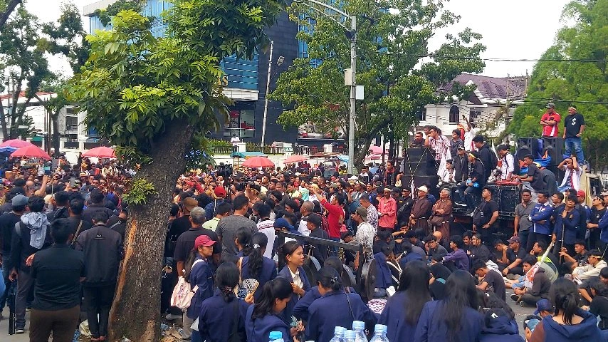 Kerumunan besar orang, termasuk mahasiswa dan masyarakat, berkumpul di luar ruangan di antara pepohonan dan bangunan untuk melakukan demonstrasi, dengan beberapa orang berdiri di atas panggung darurat atau truk di latar belakang kanan.