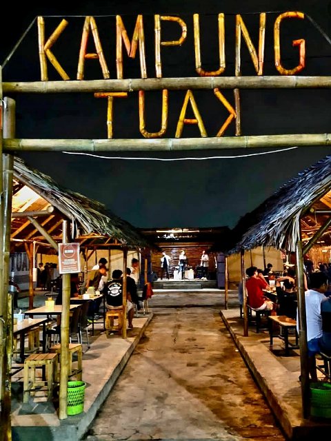 Pintu masuk Kampung Tuak Jalan Bahagia Medan dengan papan nama bambu dan suasana pengunjung menikmati tuak serta live music Batak pada malam hari.