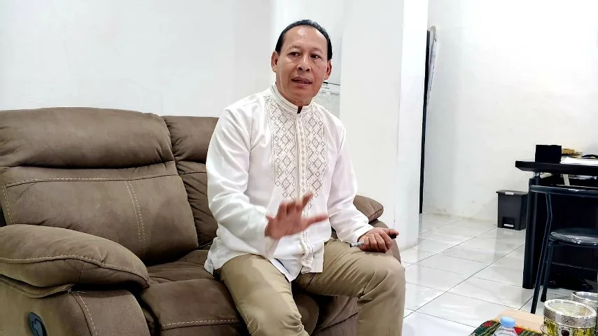 Kepala SMAN 3 Medan, Susianto, mengenakan kemeja putih duduk di sofa sambil berbicara dan menggerakkan tangan di dalam sebuah ruangan.